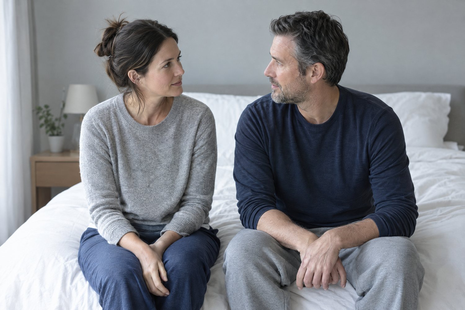 A woman and a man sitting side by side on the edge of a bed, turned toward each other and talking calmly.